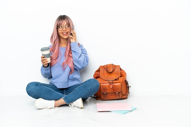 Junge Studentin gemischter Abstammung mit rosa Haaren sitzt auf dem Boden isoliert auf weißem Hintergrund und hält Kaffee zum Mitnehmen und ein Handy