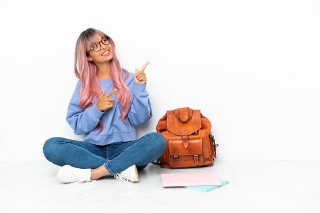 Junge Studentin gemischter Abstammung mit rosa Haaren sitzt auf dem Boden isoliert auf weißem Hintergrund und zeigt mit dem Zeigefinger eine großartige Idee