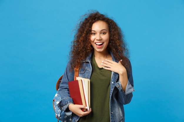 Junge Studentin in Jeanskleidung und Rucksack hält Bücher isoliert auf blauer Wand