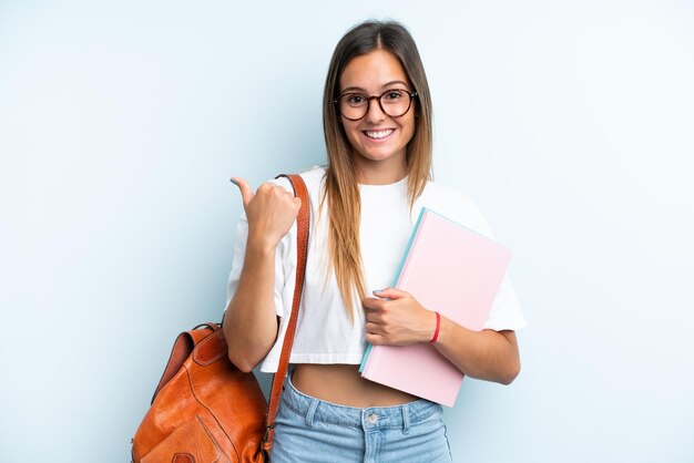 Junge Studentin isoliert auf blauem Hintergrund, die zur Seite zeigt, um ein Produkt zu präsentieren