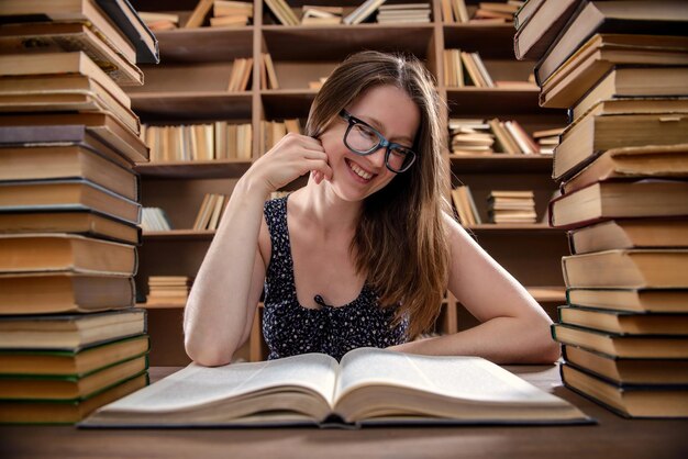 Junge Studentin liest ein Lehrbuch, das an einem Tisch mit vielen Büchern in der Bibliothek sitzt