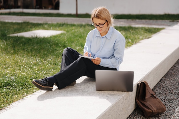 Junge Studentin mit blonden Haaren in Brillen und lässigem Outfit, die am Laptop an der frischen Luft studiert. Konzept der modernen Technologie und des urbanen Lebensstils.