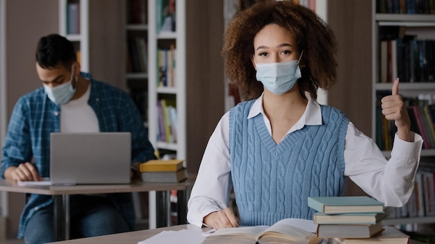 Junge Studentin mit medizinischer Maske sitzt am Schreibtisch in der Bibliothek und bereitet sich auf den Unterricht vor, in dem sie Notizen schreibt