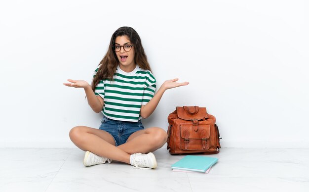 Junge Studentin sitzt auf dem Boden isoliert auf weißem Hintergrund mit schockiertem Gesichtsausdruck