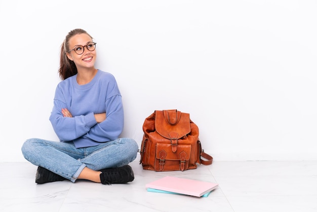 Junge Studentin sitzt auf dem Boden isoliert auf weißem Hintergrund mit verschränkten Armen und glücklich