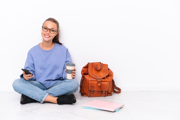 Junge Studentin sitzt auf dem Boden isoliert auf weißem Hintergrund und hält Kaffee zum Mitnehmen und ein Handy