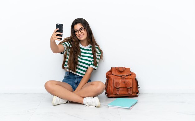 Junge Studentin sitzt auf dem Boden isoliert auf weißem Hintergrund und macht ein Selfie