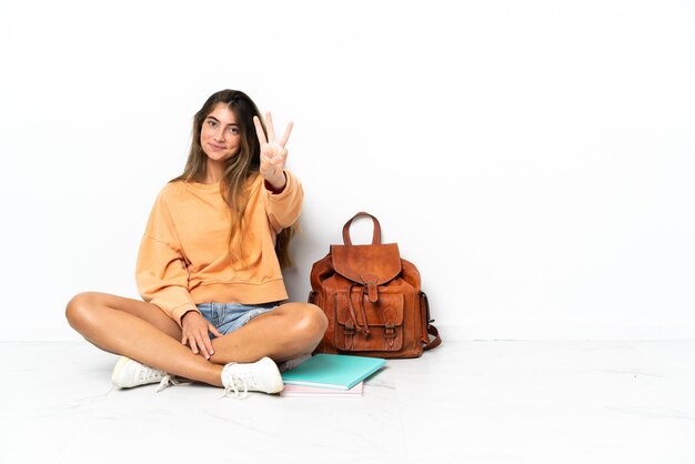 Junge Studentin sitzt auf dem Boden mit einem Laptop lokalisiert auf weißem Hintergrund glücklich und zählt drei mit den Fingern