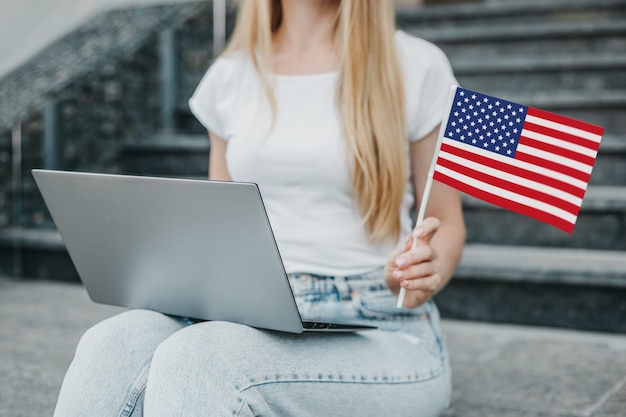 Junge Studentin sitzt auf Treppen, zeigt eine kleine amerikanische Flagge im Hintergrund der Universität. Nahaufnahme