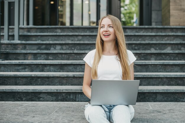 Junge Studentin sitzt mit Laptop auf der Treppe in der Nähe der Universität, lächelt und arbeitet and