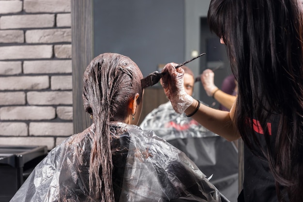 Junge Stylistin steht und beendet den Prozess des Färbens der Haare einer jungen Frau, während sie im Friseursessel im Schönheitssalon sitzt