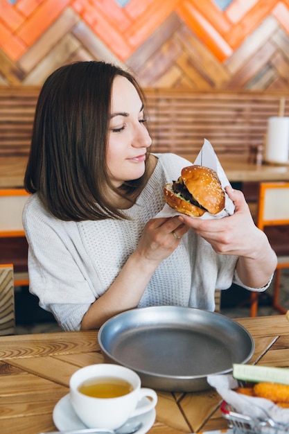 Junge, süße, brünette Frau, die im Café einen frisch zubereiteten leckeren Burger isst. Tonen.