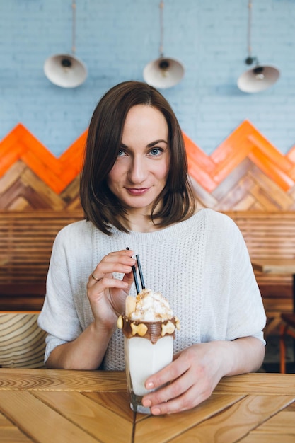 Junge, süße, brünette Frau trinkt Milchshake, der wunderschön mit Sahne und Keksen im Café dekoriert ist. Tonen.