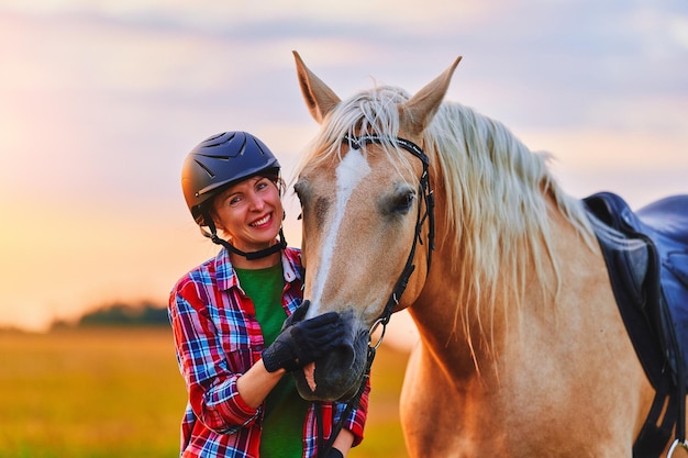 Junge, süße, fröhliche, zufrieden lächelnde Reiterin mit Helm, die zur goldenen Stunde auf der Wiese ein schönes blondes Palomino-Pferd umarmt und streichelt