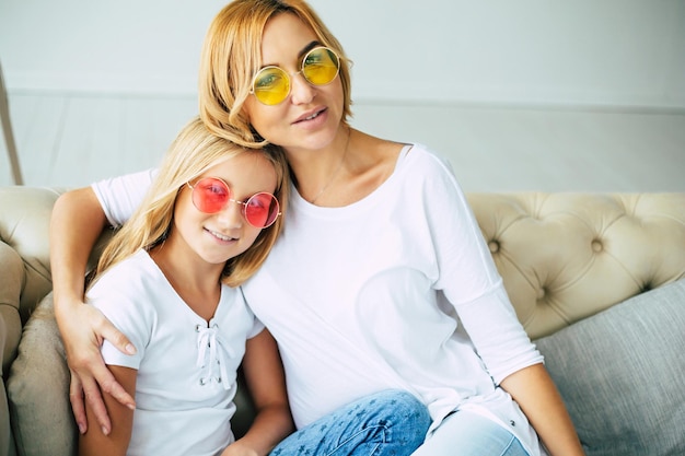 Junge süße Mutter und kleine Tochter in bunten Sonnenbrillen haben Spaß und umarmen sich auf der Couch zu Hause