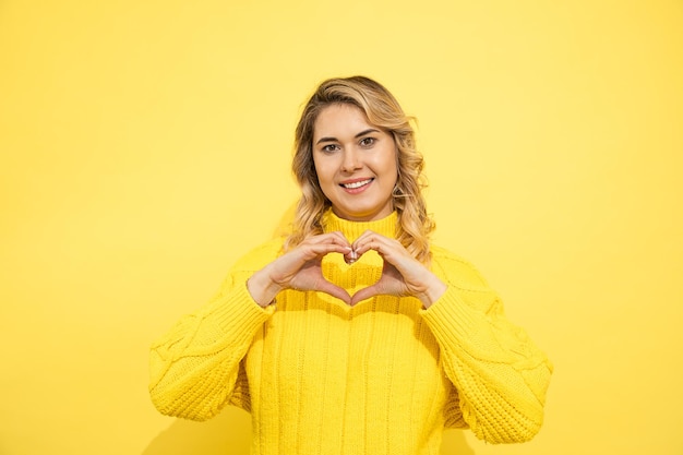 Junge süße, schöne blonde Frau mit Pullover über gelber Wand isolierter Hintergrund, der verliebt lächelt und Herzformsymbol und -form mit den Händen zeigt. Dating-Konzept. Studioaufnahme