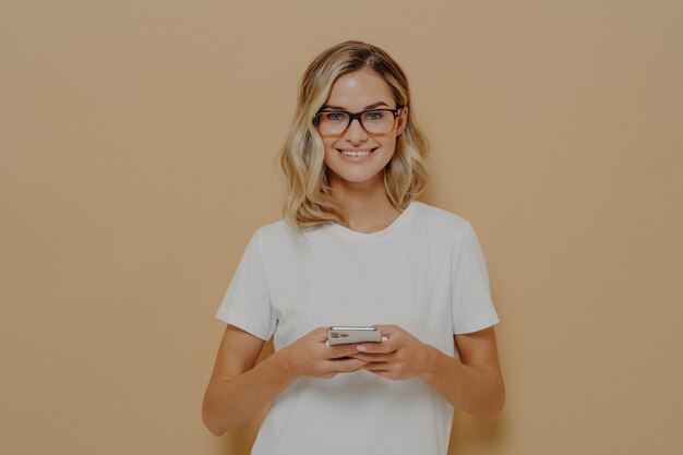 Junge süße Studentin in Brillen mit Handy und Blick in die Kamera mit angenehmem Lächeln, Tippen an Freunde oder Chatten in sozialen Medien. Moderne Technologien und Kommunikationskonzept