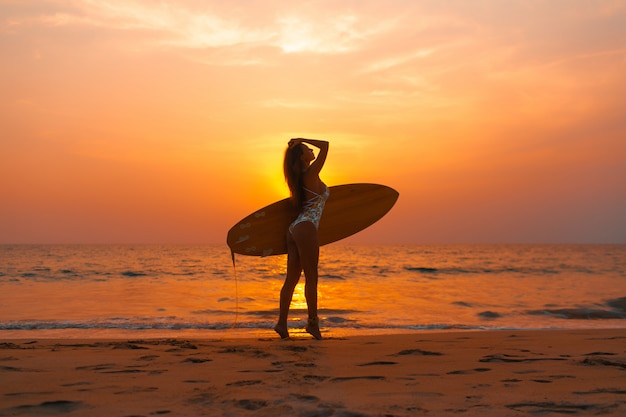 Junge Surferfrau in einem trendigen Bikini geht mit einem Surfbrett in ihren Händen am Ozeansonnenuntergang.