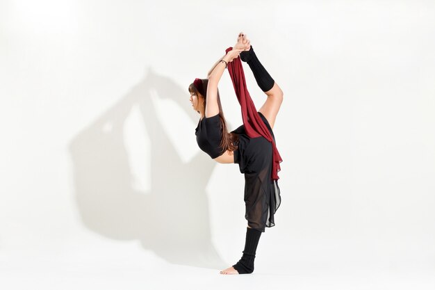 Junge Tänzerin, die eine Eislauf-Biellman-Pose durchführt, die sich nach oben streckt, um ihren Fuß zu greifen, während sie auf einem Bein in einer Seitenansicht, isoliert auf Weiß mit Schatten und Exemplar, balanciert