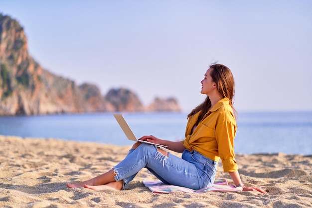 Junge tausendjährige Freiberuflerin mit Laptop am Strand auf dem Seeweg Traumbüro-Arbeitskonzept