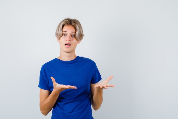 Junge Teenager, die eine Geste der Frage im blauen T-Shirt machen und verwirrt aussehen. Vorderansicht.