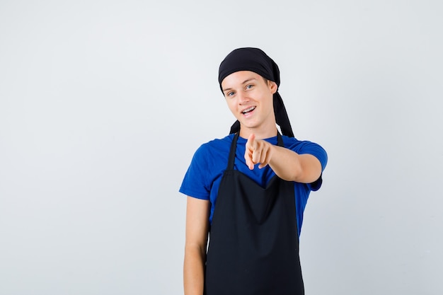 Junge Teenager-Koch im T-Shirt, Schürze nach vorne zeigend und fröhlich aussehend, Vorderansicht.