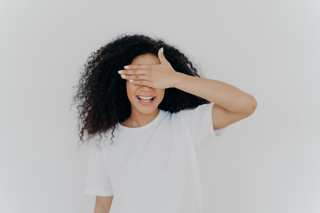 Junge Teenie-Dame bedeckt Augen mit Handfläche, versteckt sich, lächelt zahnig, hat buschige Afro-Frisur, trägt weißes Mockup-T-Shirt, steht allein im Studio, wartet auf Überraschung, aufgeregt vor Vorfreude