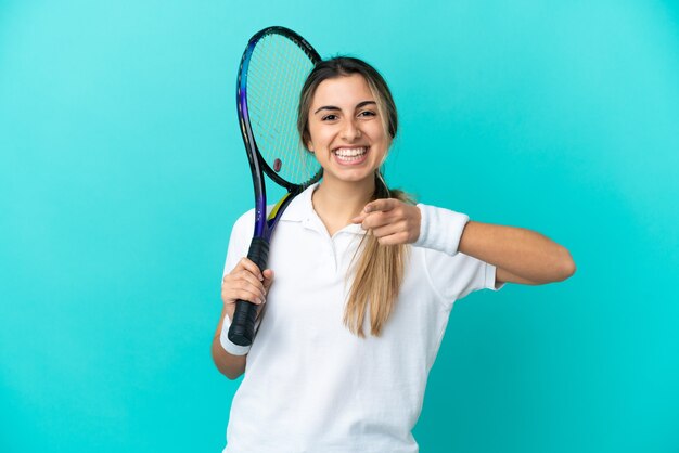 Junge Tennisspielerin lokalisiert auf blauem Hintergrund überrascht und nach vorne zeigend