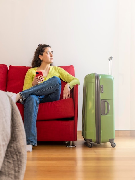 Junge Touristenfrau mit ihrem Smartphone in einem Hotelzimmer, die auf einem Sofa sitzt und aus dem Fenster schaut