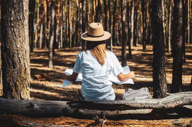 Junge Touristin in Hut und T-Shirt sitzt auf einem Baumstamm und sieht sich während eines Halts im Wald eine Karte an. Rückansicht.