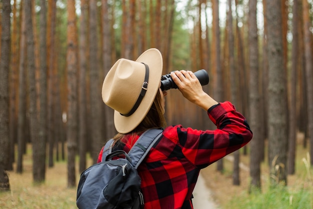 Junge Touristin mit Rucksack, Hut und rotem kariertem Hemd und schaut durch ein Fernglas