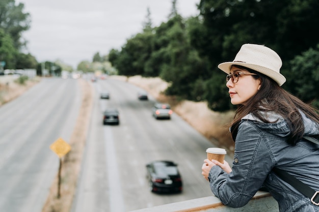junge Touristin mit Strohhut, die auf einer Fußgängerüberführung steht und eine Tasse Kaffee hält, blickt auf die Autobahn mit Autos, die an einem sonnigen Tag durchfahren. weibliche reiserucksacktouristin lächelnd im freien.