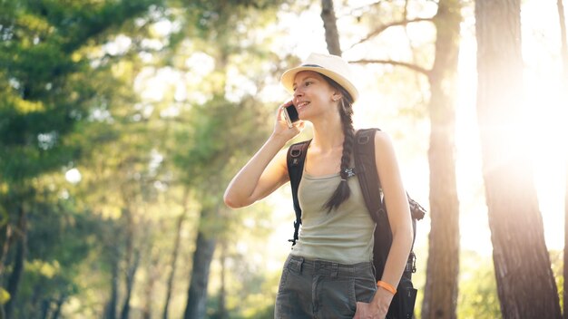 Junge touristische Rucksacktouristin mit Hut, die beim Wandern im Sommerwald mit dem Smartphone plaudert