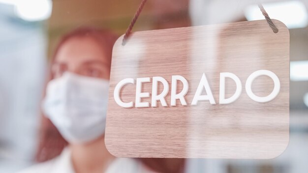 Junge traurige Frau mit Gesichtsmaske ändert das Schild Offen zu geschlossen auf Spanisch auf dem Fenster für das Corona-Virus