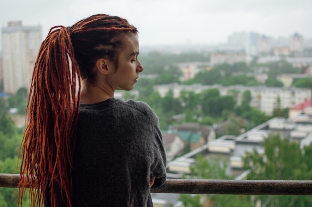 Junge traurige Frau mit roten Dreadlocks blickt nach dem Regen vom Balkon auf die Stadt und wartet.