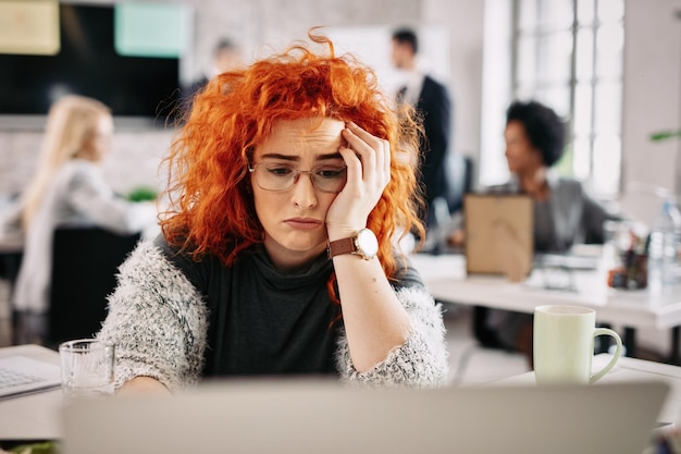 Junge traurige Geschäftsfrau, die im Büro arbeitet und an etwas denkt Es gibt Leute im Hintergrund