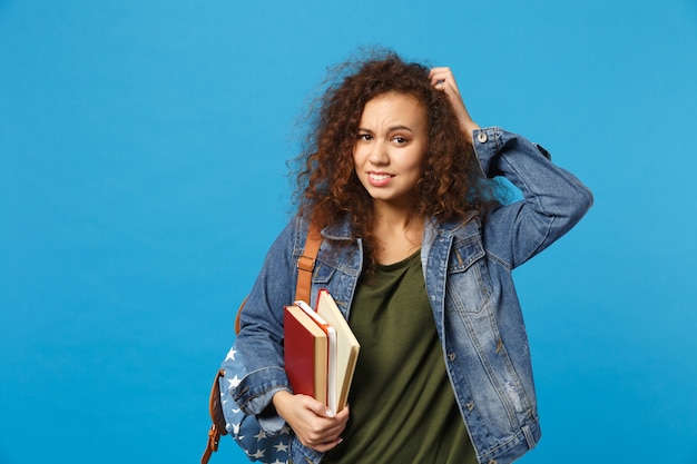 Junge traurige Studentin in Jeanskleidung und Rucksack hält Bücher isoliert auf blauer Wand