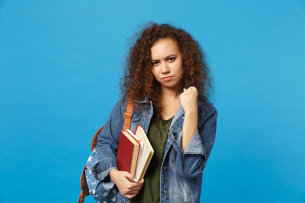 Junge traurige Studentin in Jeanskleidung und Rucksack hält Bücher isoliert auf blauer Wand
