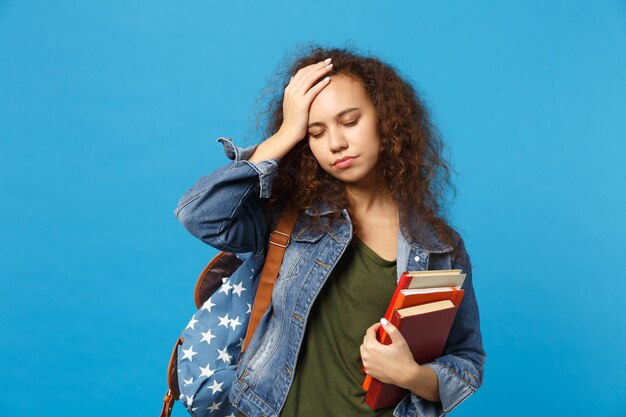 Junge traurige Studentin in Jeanskleidung und Rucksack hält Bücher isoliert auf blauer Wand