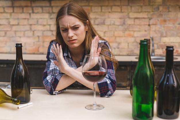 Junge traurige und verschwendete alkoholische Frau, die an der Küchencouch sitzt und Rotwein trinkt, völlig betrunken, einsam deprimiert und unter Kater in Alkoholismus und Alkoholmissbrauch leidet.