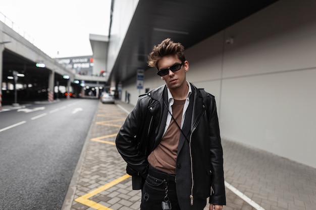 Junge trendige gutaussehende Mann Hipster mit Sonnenbrille mit Frisur in einem Vintage-Hemd in einer stilvollen schwarzen Lederjacke in modischen Jeans geht auf einer Straße in der Stadt. Attraktives Kerlmodell draußen.