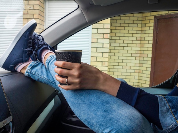 Junge trinken Kaffee zum Mitnehmen mit Füßen in Stiefeln auf dem Armaturenbrett des Autos Reisereise