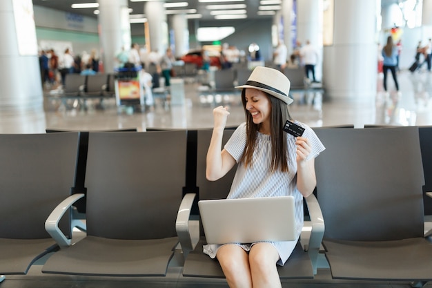 Junge überglückliche reisende Touristenfrau, die am Laptop arbeitet, Kreditkarte hält, Gewinnergeste macht, in der Lobbyhalle am Flughafen wartet?