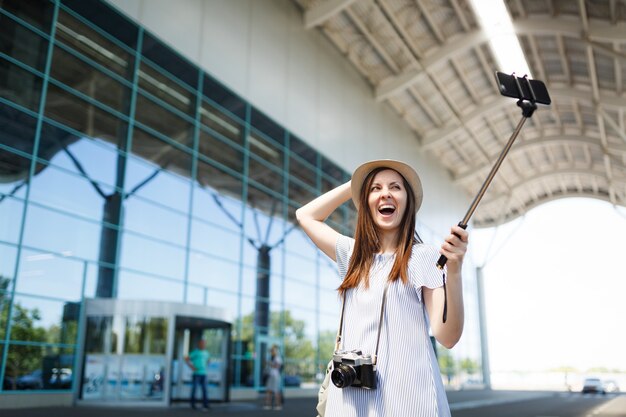 Junge überglückliche reisende Touristenfrau mit Retro-Vintage-Fotokamera, die Selfie auf dem Handy mit egoistischem Einbeinstativ am Flughafen macht?