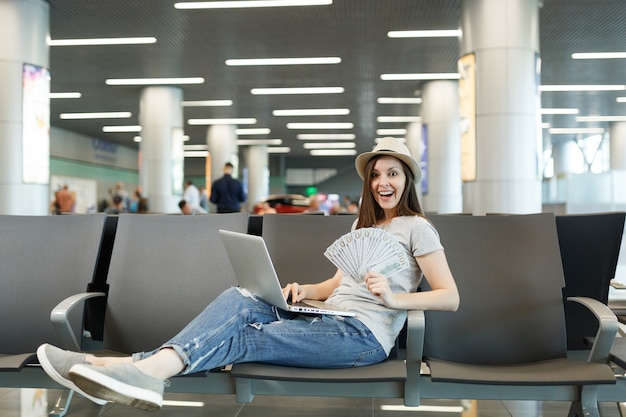 Junge überraschte reisende Touristenfrau, die am Laptop arbeitet, hält Bündel von Dollar-Bargeld in der Lobbyhalle am internationalen Flughafen?