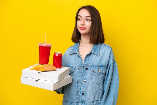 Junge ukrainische Frau, die Fast Food isoliert auf gelbem Hintergrund hält, zur Seite schaut und lächelt