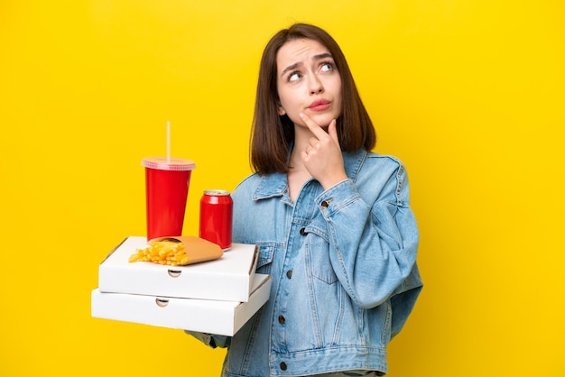 Junge ukrainische Frau, die Fastfood isoliert auf gelbem Hintergrund hält und Zweifel hat