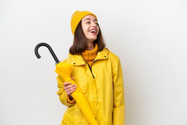 Junge ukrainische Frau mit regenfestem Mantel und Regenschirm isoliert auf weißem Hintergrund lachend in Seitenlage