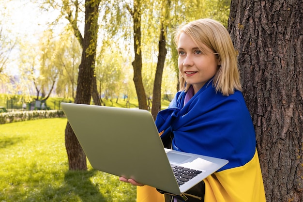 Junge ukrainische Frau mit ukrainischer Flagge benutzt einen Laptop im Freien im Park