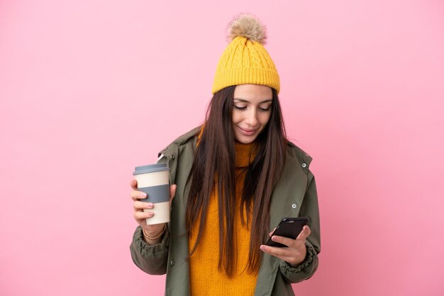 Junge ukrainische Frau mit Winterjacke isoliert auf rosa Hintergrund mit Kaffee zum Mitnehmen und einem Handy